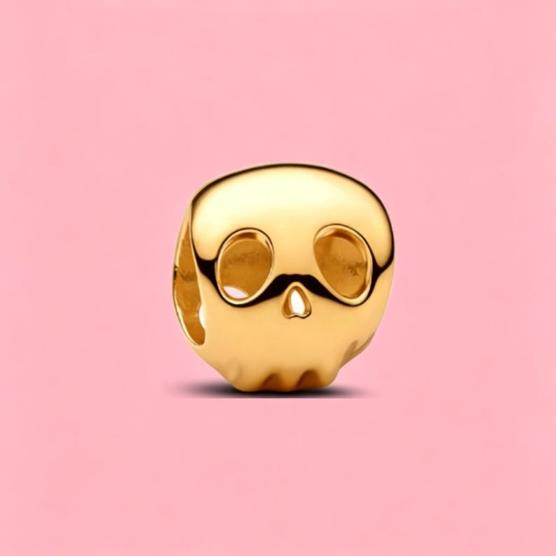 May include: A polished gold-tone metal bead shaped like a stylized skull. The bead features smooth, rounded contours with two large, hollowed-out eye sockets and a small, triangular nasal cavity, designed with a central hole for threading.