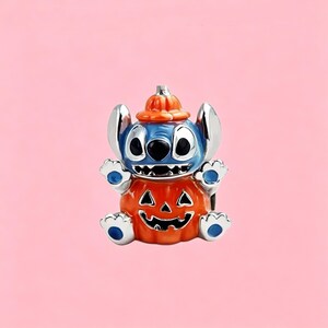 May include: A silver-toned metal charm featuring a blue character with large ears wearing an orange pumpkin hat and sitting inside a carved orange jack-o'-lantern pumpkin with a black face design, featuring blue accents on the hands and feet.