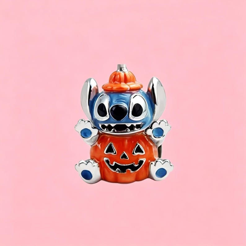 May include: A silver-toned metal charm featuring a blue character with large ears wearing an orange pumpkin hat and sitting inside a carved orange jack-o'-lantern pumpkin with a black face design, featuring blue accents on the hands and feet.