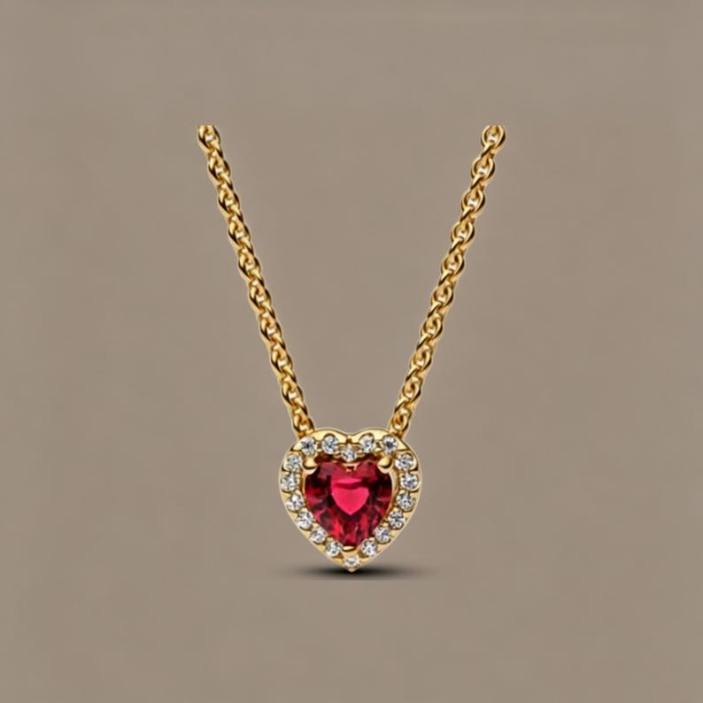 May include: A gold chain necklace featuring a heart-shaped pendant with a central red gemstone surrounded by a halo of small clear stones. The pendant is suspended from a delicate gold link chain, creating a classic and elegant jewellery piece.