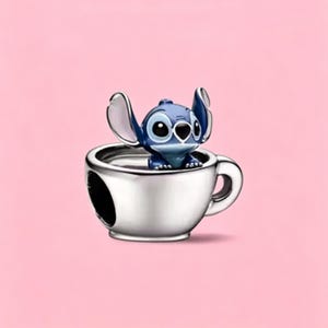 May include: A silver-toned metal charm shaped like a teacup with a handle, featuring a small, blue, cartoon-style creature with large ears and dark eyes sitting inside the cup. The charm has a smooth, polished finish and a hollow center.