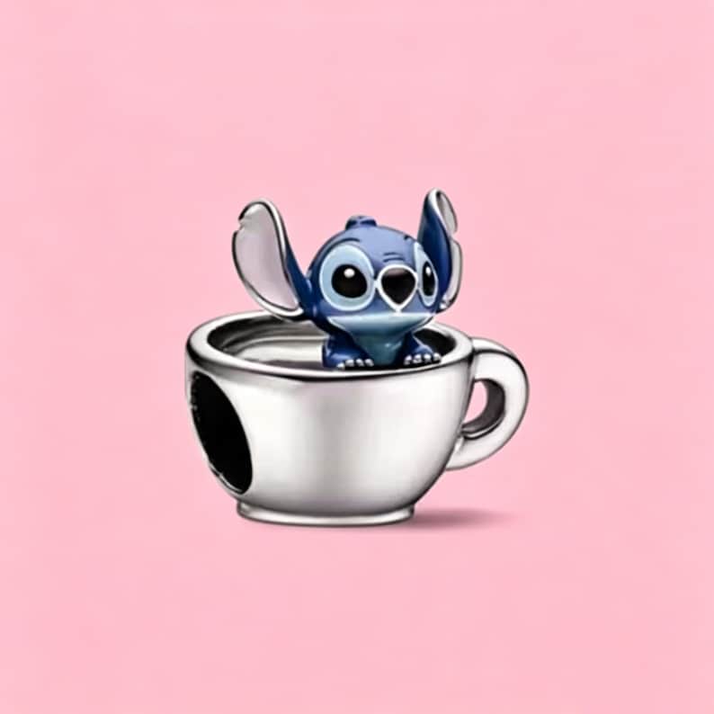May include: A silver-toned metal charm shaped like a teacup with a handle, featuring a small, blue, cartoon-style creature with large ears and dark eyes sitting inside the cup. The charm has a smooth, polished finish and a hollow center.