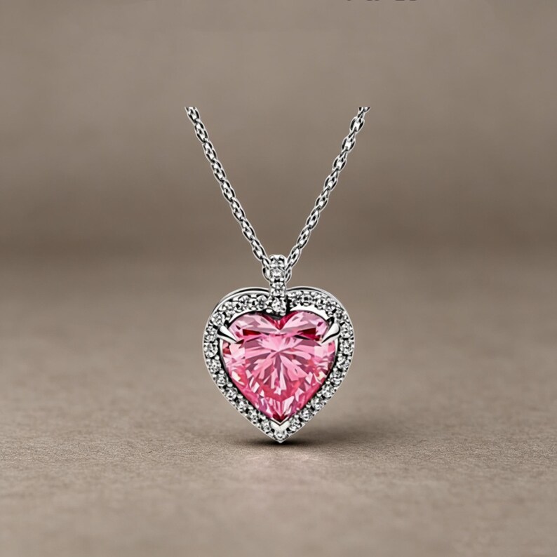 May include: A silver-toned necklace featuring a heart-shaped pendant with a large, faceted pink centre stone. The heart is surrounded by a halo of small, clear, sparkling stones set in metal, suspended from a delicate silver-toned chain.