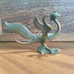 Pode incluir: Uma estatueta metálica em tom de bronze de uma ave mítica com pescoço alongado, uma crista na cabeça e asas grandes e abertas. A criatura assenta sobre uma base com padrões de redemoinho e apresenta um acabamento com pátina envelhecida.