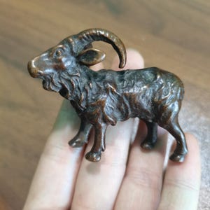 May include: A small, dark bronze-coloured metal figurine of a mountain goat with curved horns, a textured coat, and a beard. The sculpture is depicted in a standing position, showcasing detailed facial features and muscular legs in a compact size.