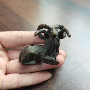 May include: A small, dark bronze-toned metal figurine of a ram in a resting position. The sculpture features detailed textures representing fur on the body and neck, along with prominent, curved, spiralling horns extending from the head.