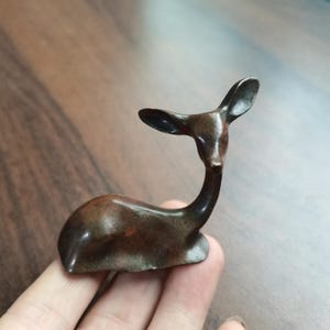 May include: A small, dark brown metallic figurine of a deer in a resting position. The deer has a smooth, curved body, a slender neck, and upright ears, with a polished finish that shows subtle variations in the dark brown metal coloration.