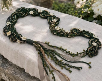 Op de afbeelding: Een decoratieve gevlochten slinger met donkergroen en goudkleurig koord in een cirkelvormig patroon, geaccentueerd met kleine witte bloemen. Het stuk bevat hangende linten, gevlochten strengen en verfijnde groene bladversieringen.