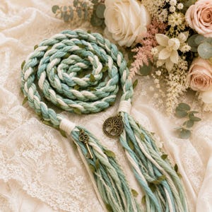 May include: A braided cord in shades of sage green, teal, and cream, interwoven with small green leaves. The cord features a bronze-toned metal charm of a tree of life and a vintage-style skeleton key charm, secured with white thread wrappings.