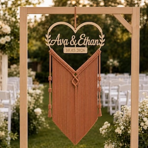 May include: A heart-shaped wooden sign featuring the laser-cut names Ava & Ethan and the date 10.03.2026, suspended above a terracotta-colored macrame wall hanging with intricate knotted patterns and long, straight fringe hanging in a V-shape.