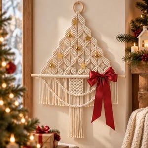 May include: A cream-colored macrame wall hanging shaped like a Christmas tree, featuring a wooden ring at the top, a horizontal wooden dowel, gold star ornaments, small gold spheres, and a large, shiny red satin ribbon bow tied on the right.