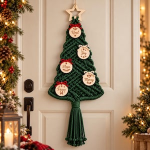 May include: A dark green macrame wall hanging shaped like a Christmas tree, topped with a wooden star. It features five round wooden ornaments with the words Noel, Joy, Hope, Merry, and Peace, accented with small red and white fabric bows.
