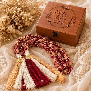Puede incluir: Una caja rectangular de madera con cierre metálico, grabada con el texto Emily & Lucas y May 24, 2025, dentro de un diseño de corona floral. Junto a ella hay un cordón trenzado con hilos rojo oscuro, crema y dorados con borlas a juego.