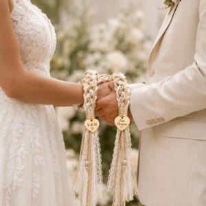 Pode incluir: Um cordão de união de mãos em corda trançada creme e dourada, adornado com dois pingentes dourados em forma de coração. Um pingente tem gravado K & M e um coração, o outro diz Our Forever com um coração. O cordão termina em borlas de renda.
