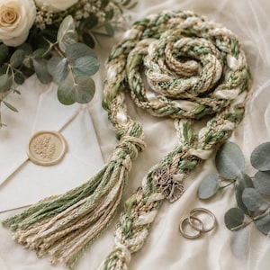 Puede incluir: Un cordón trenzado hecho de fibras entrelazadas en color verde salvia, crema y beige, con una borla grande en un extremo. El cordón está adornado con un dije de árbol plateado y dos dijes de corazón entrelazados, junto a dos anillos plateados.
