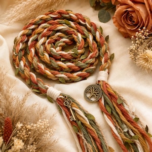 May include: A braided cord in shades of rust orange, olive green, and cream, interwoven with small green leaves. The cord features a bronze-toned metal tree of life charm and a vintage-style skeleton key charm attached to the ends with cream thread.