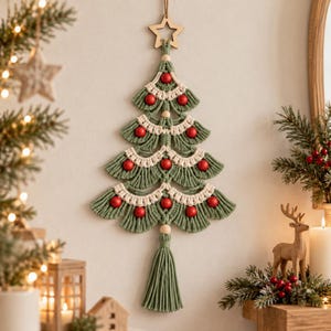 May include: A macrame wall hanging shaped like a Christmas tree, crafted from sage green and cream-colored cord. It features four tiered layers decorated with smooth red wooden beads and is topped with a light-colored wooden star ornament.