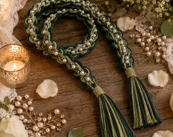 May include: A braided cord belt in dark green and sage green yarn, embellished with white faux pearls throughout the length. The belt features two long tassels at the ends, each wrapped with matching green thread, creating a decorative accessory.