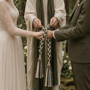 May include: A handfasting cord featuring braided cream and dark green rope intertwined with delicate green leaf vines. The ends are finished with cream-colored thread bindings and circular wooden charms depicting a tree of life motif.