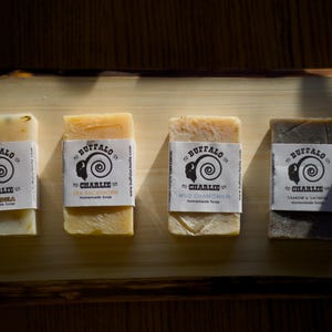 May include: Four bars of handmade soap with a black and white logo featuring a buffalo head and the text "Buffalo Charlie" on each bar. The soap bars are labelled with different scents: "Chamomile", "Sea Buckthorn", "Wild Chamomile", and "Chamomile & Oatmeal".