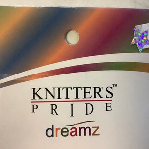 Puede incluir: Un paquete blanco con un fondo degradado de colores. El paquete tiene un agujero en la parte superior y una pegatina de estrella holográfica. El texto "KNITTERS' PRIDE" está impreso en negro con un subrayado rojo. El texto "dreamz" está impreso en colores arcoíris.