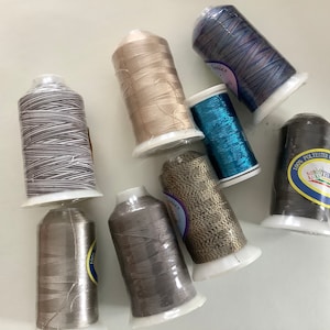 May include: Assortment of eight spools of thread in various colors, including silver, gray, brown, blue, and iridescent. The spools are wrapped in clear plastic and have labels with text.