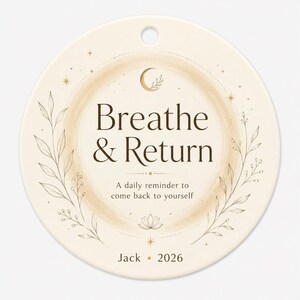 May include: A round cream-colored ceramic ornament featuring a hole at the top. The text reads "Breathe & Return", "A daily reminder to come back to yourself", "Jack", and "2026". It includes gold-toned illustrations of a crescent moon, leaves, and stars.