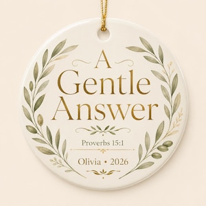 May include: A round white ceramic ornament featuring gold-coloured text that reads A Gentle Answer, Proverbs 15:1, Olivia, and 2026. The design is framed by a watercolour-style olive branch wreath in shades of green, suspended by a gold string.