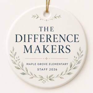 May include: A round white ceramic ornament featuring a gold hanging string. The text reads THE DIFFERENCE MAKERS, MAPLE GROVE ELEMENTARY, and STAFF 2026 in dark blue font. A delicate watercolor illustration of green leaves frames the bottom text.