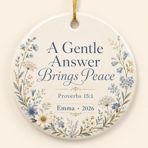 May include: A round ceramic ornament featuring a gold hanging string. The centre displays the dark blue text A Gentle Answer Brings Peace, Proverbs 15:1, Emma, and 2026. The design is framed by a delicate watercolour illustration of blue and white wildflowers.