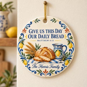 May include: A round ceramic wall hanging featuring the text Give Us This Day Our Daily Bread Matthew 6:11 and The Harris Family. The design includes a loaf of bread, wheat stalks, a blue and white pitcher, and yellow lemons with green leaves.