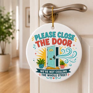 Op de afbeelding: Een rond wit keramisch hangbord met een gouden koord. De tekst luidt PLEASE CLOSE THE DOOR en WE'RE NOT COOLING THE WHOLE STREET. Het bevat een afbeelding van een open lichtblauwe deur met windlijnen en twee potplanten.