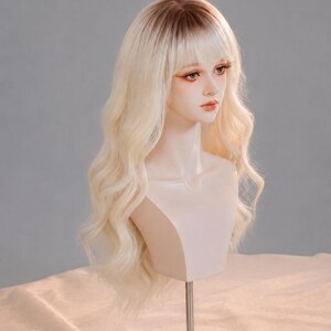May include: A long, wavy synthetic wig featuring a light blonde color with dark brown roots and straight bangs. The wig is displayed on a pale, neutral-toned mannequin head with painted facial features, including defined eyes and lips, set on a stand.