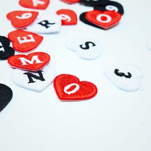 Puede incluir: Una colección de parches bordados en forma de corazón en rojo, blanco y negro. Cada parche presenta una letra o número bordado en un color contrastante. Los parches están dispersos, mostrando varios caracteres que incluyen M, N, O, R, S, E y números.