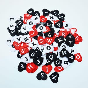 Puede incluir: Una colección de pequeños parches bordados en forma de corazón en rojo, blanco y negro. Cada parche presenta una letra o número bordado en un color contrastante. Los parches están esparcidos en un montón, mostrando varios caracteres.