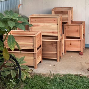 May include: A collection of rectangular wooden planters constructed from light brown cedar planks. The planters feature a slatted design with vertical corner posts and open tops, arranged in a stacked and grouped configuration on a concrete surface.