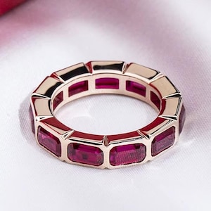 Könnte beinhalten: Ein Ring aus Roségold mit einer durchgehenden Reihe rechteckiger, tiefrosa facettierter Edelsteine in einer Kanalfassung. Der Ring hat eine polierte metallische Oberfläche und ein einheitliches Design, das den gesamten Umfang des Bandes umschließt.