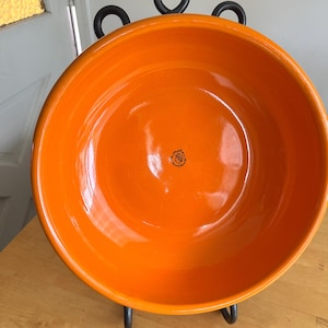 May include: A bright orange ceramic bowl with a smooth, glossy finish. The interior features a small, centered black stamp depicting a profile silhouette. The bowl is supported by a black metal stand with three circular loops at the top.