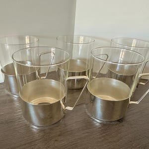 May include: A set of five clear glass cups, each featuring a removable silver-toned metal base and a matching silver-toned metal handle. The cups have a cylindrical shape with a smooth rim and a polished metallic finish on the lower exterior.