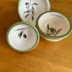 May include: Three white ceramic bowls with green rims. The largest bowl features a painted olive branch and the text Provença. The medium bowl displays a green olive branch and the text Cerignola. The smallest bowl shows a single dark olive branch.