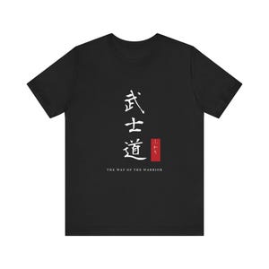 May include: A black short-sleeved t-shirt featuring white Japanese kanji characters vertically aligned in the center. Below the characters is a small red rectangular graphic and the white text THE WAY OF THE WARRIOR printed underneath.