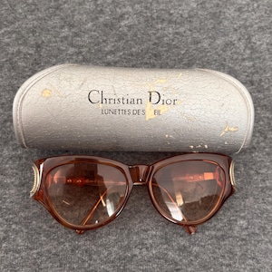 May include: Brown sunglasses with a rounded frame and gold-tone side accents, paired with a silver-coloured hard case featuring the text Christian Dior LUNETTES DE SOLEIL. The case has a textured finish with visible signs of wear and surface peeling.