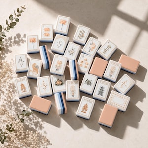 May include: A collection of rectangular mahjong tiles featuring white faces with intricate illustrations of flowers, pagodas, and the word Joker, alongside solid peach-colored tiles and blue-striped tiles, all arranged on a neutral surface.