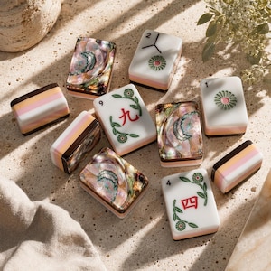May include: A collection of rectangular mahjong tiles featuring a mix of white tiles with green and red floral designs and Chinese characters, alongside tiles with iridescent, multi-colored abalone shell inlays and layered pastel-colored bases.