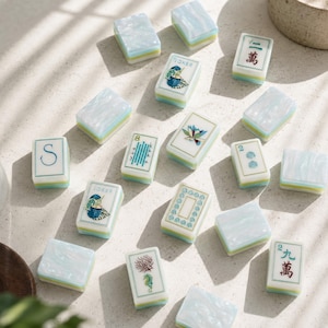 May include: A collection of rectangular mahjong tiles featuring a mix of solid pearlescent light blue surfaces and tiles decorated with various illustrations including a cocktail, a lotus flower, a seahorse, Chinese characters, and the letter S.