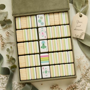 May include: A set of rectangular mahjong tiles arranged in a green velvet-lined box. The tiles feature a light wood grain finish with vertical light green stripes. Several tiles display colorful floral and numerical symbols in pink, green, and blue.