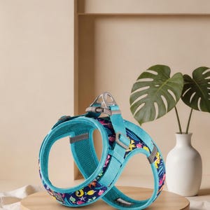 May include: A teal and dark blue pet harness featuring a colourful pattern of sea creatures including fish, turtles, and starfish. The harness includes adjustable straps, a metallic D-ring for leash attachment, and a breathable mesh interior lining.