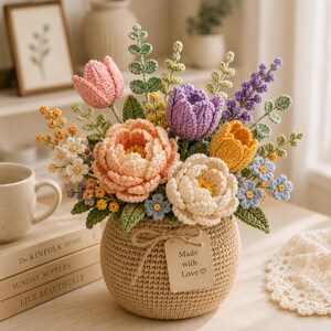 Könnte beinhalten: Ein gehäkeltes Blumenarrangement in einem runden beigen Topf mit einem Etikett mit der Aufschrift Made with Love. Der Strauß besteht aus rosa, lila und gelben Tulpen, pfirsichfarbenen und cremefarbenen Pfingstrosen, kleinen blauen und weißen Blüten.