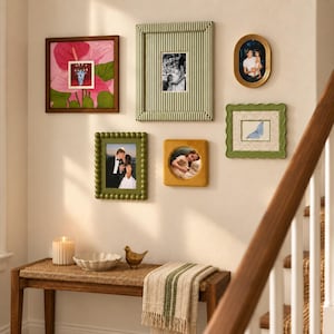 May include: A gallery wall arrangement featuring six diverse picture frames, including a square wooden frame with a pink floral print, a green striped rectangular frame, an oval gold frame, a scalloped green frame, a beaded green frame, and a mustard yellow square frame with a circular opening.