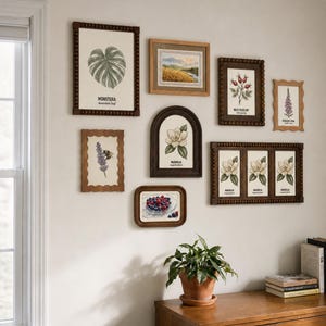 May include: A gallery wall arrangement featuring eight framed botanical and nature prints. The collection includes illustrations of a monstera leaf, a landscape, wild rose hips, foxglove, lavender with a bee, a magnolia, berries, and three magnolia flowers.